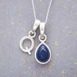 Sterling silver 'Q' initial necklace with a September birthstone blue sapphire pear-shaped pendant on a grey background.