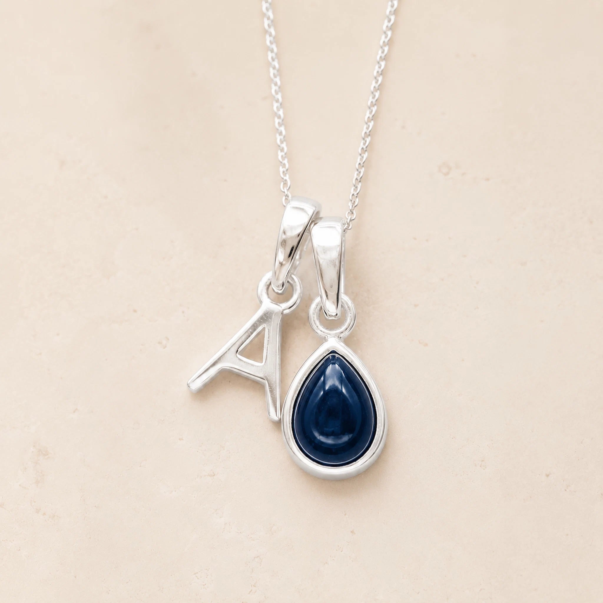 Sterling silver 'A' initial necklace with a September birthstone blue sapphire pear-shaped pendant on a grey background.