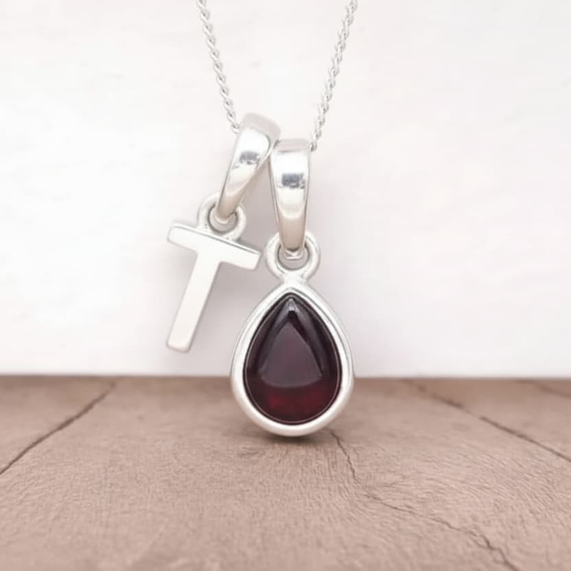 Silver Initial T necklace with a teardrop-shaped Garnet gemstone pendant hanging above a rustic wooden surface