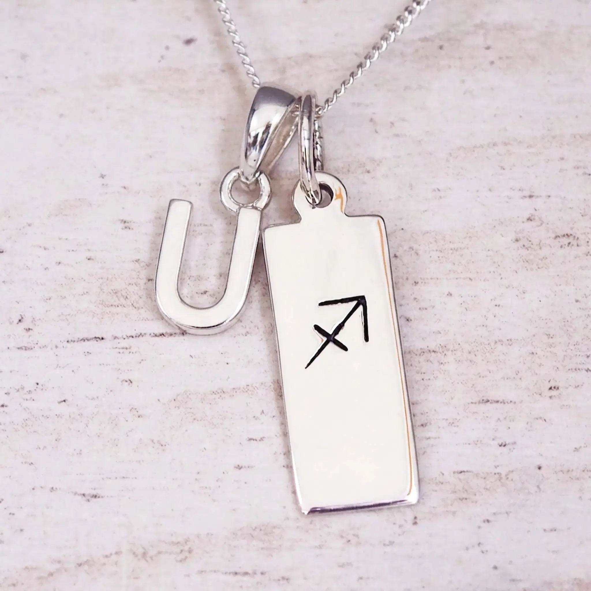 Sterling silver initial U and Sagittarius zodiac bar necklace, close-up of the letter charm and pendant on whitewashed wood