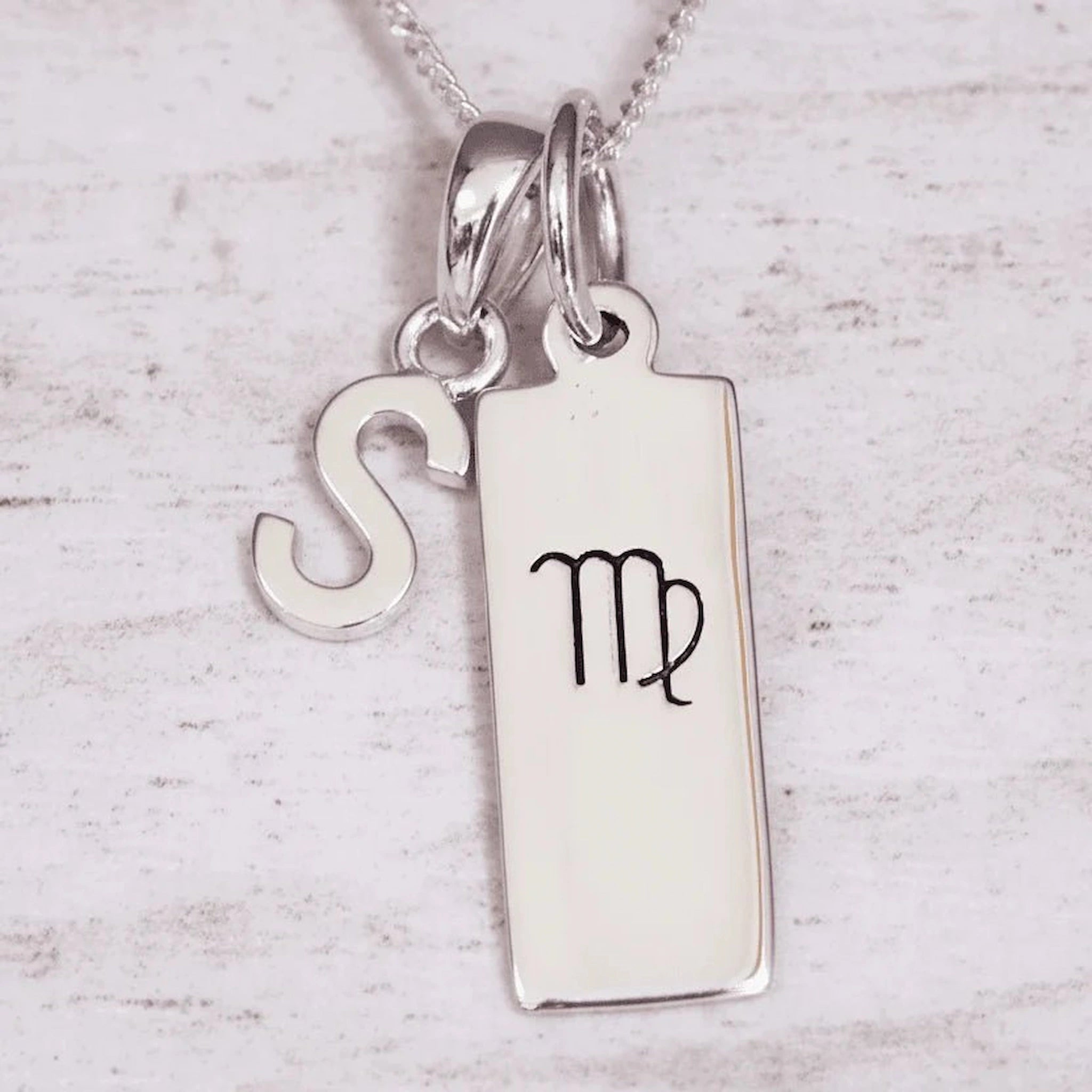 sterling Silver double pendant necklace with a rectangle pendant with the virgo zodiac glyph, and a 'S' initial pendant on a light wooden background