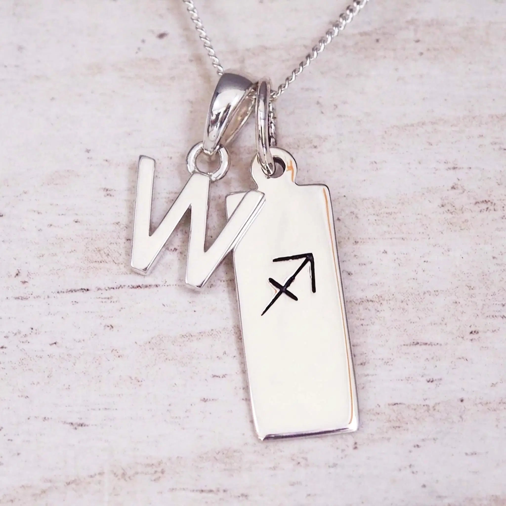 Sterling silver initial W and Sagittarius zodiac bar necklace, close-up of the letter charm and pendant on whitewashed wood