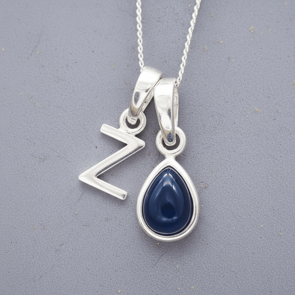 Two silver pendants on a chain against a grey background, a uppercase Z pendant and a pear shaped september birthstone sapphire pendant.