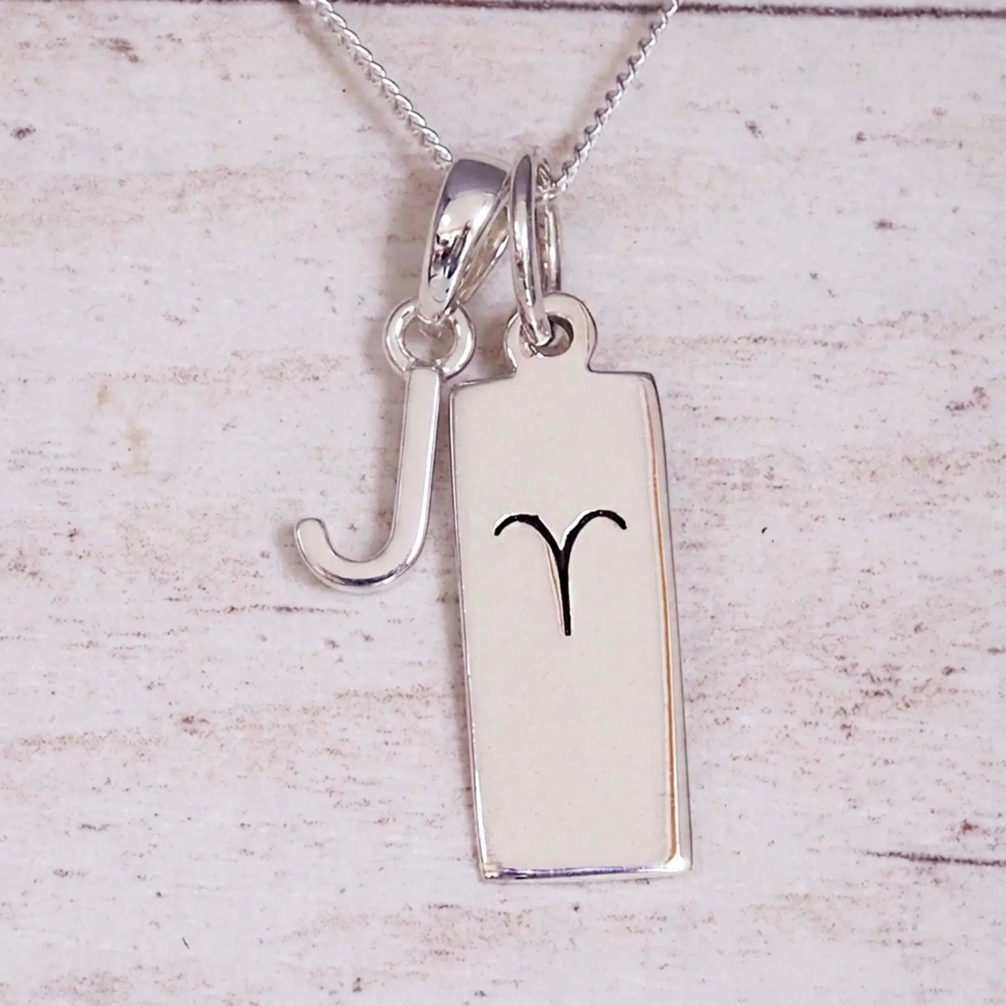 Sterling Silver J Initial pendant and Aries necklace on a fine silver chain on a light wooden background