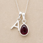 Silver necklace with 'A' pendant and teardrop-shaped garnet stone on beige background