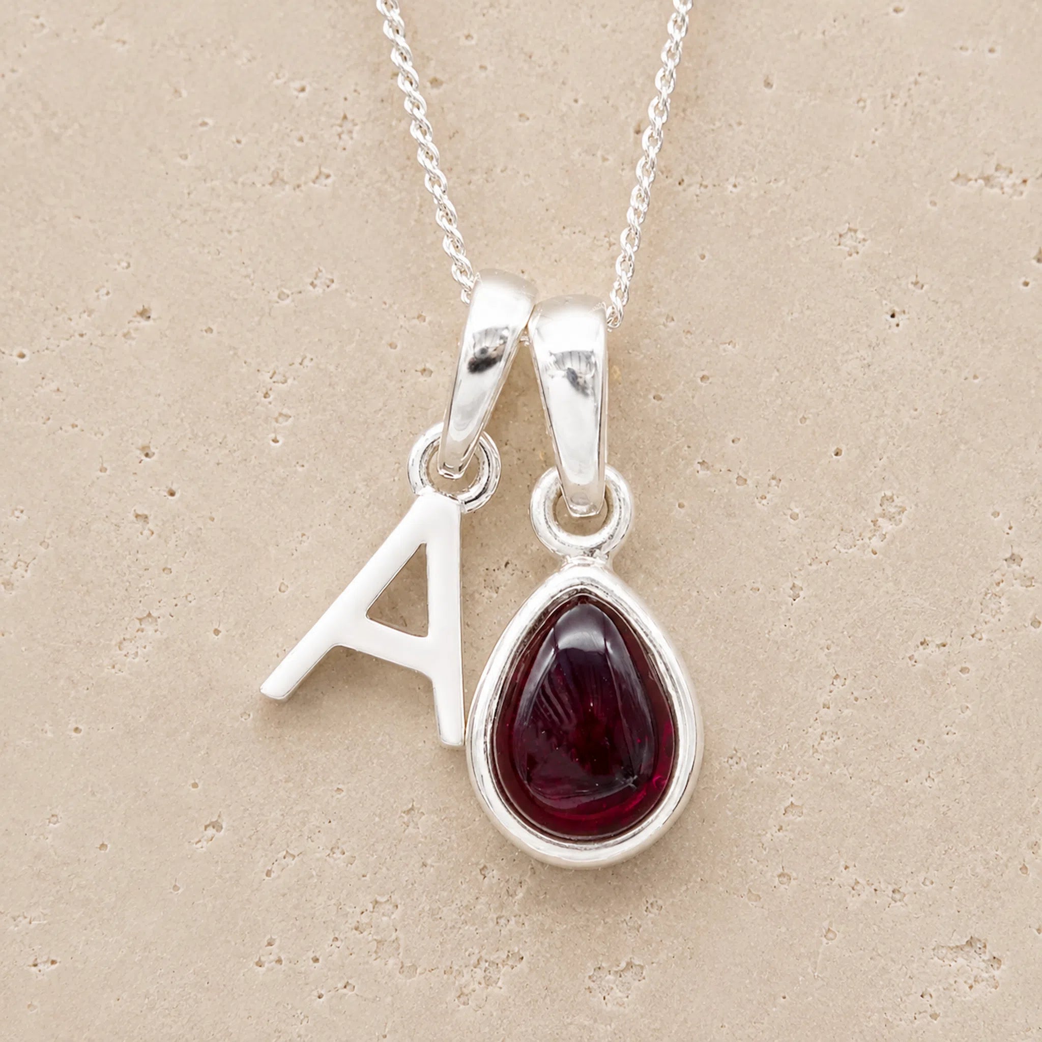 Silver necklace with 'A' pendant and teardrop-shaped garnet stone on beige background