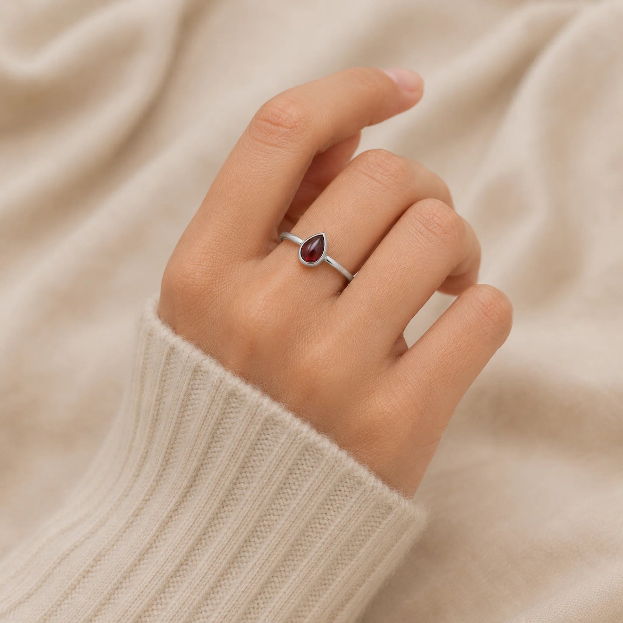 Hand wearing a silver ring with a red gemstone on a beige fabric background