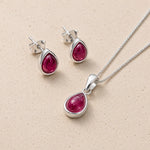 Silver july birthstone jewellery set featuring a teardrop-shaped ruby pendant necklace and matching stud earrings, displayed on a textured beige background.