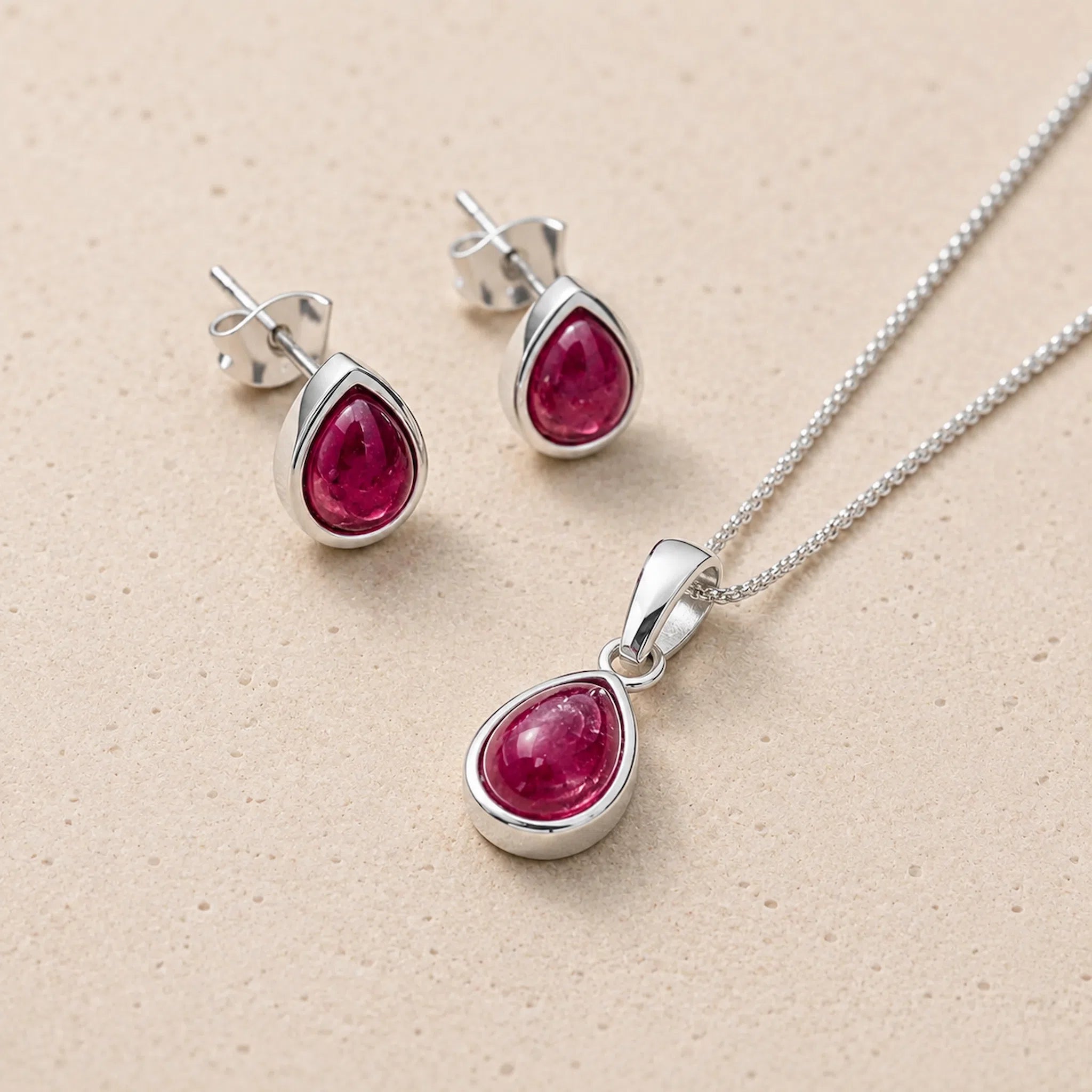 Silver july birthstone jewellery set featuring a teardrop-shaped ruby pendant necklace and matching stud earrings, displayed on a textured beige background.