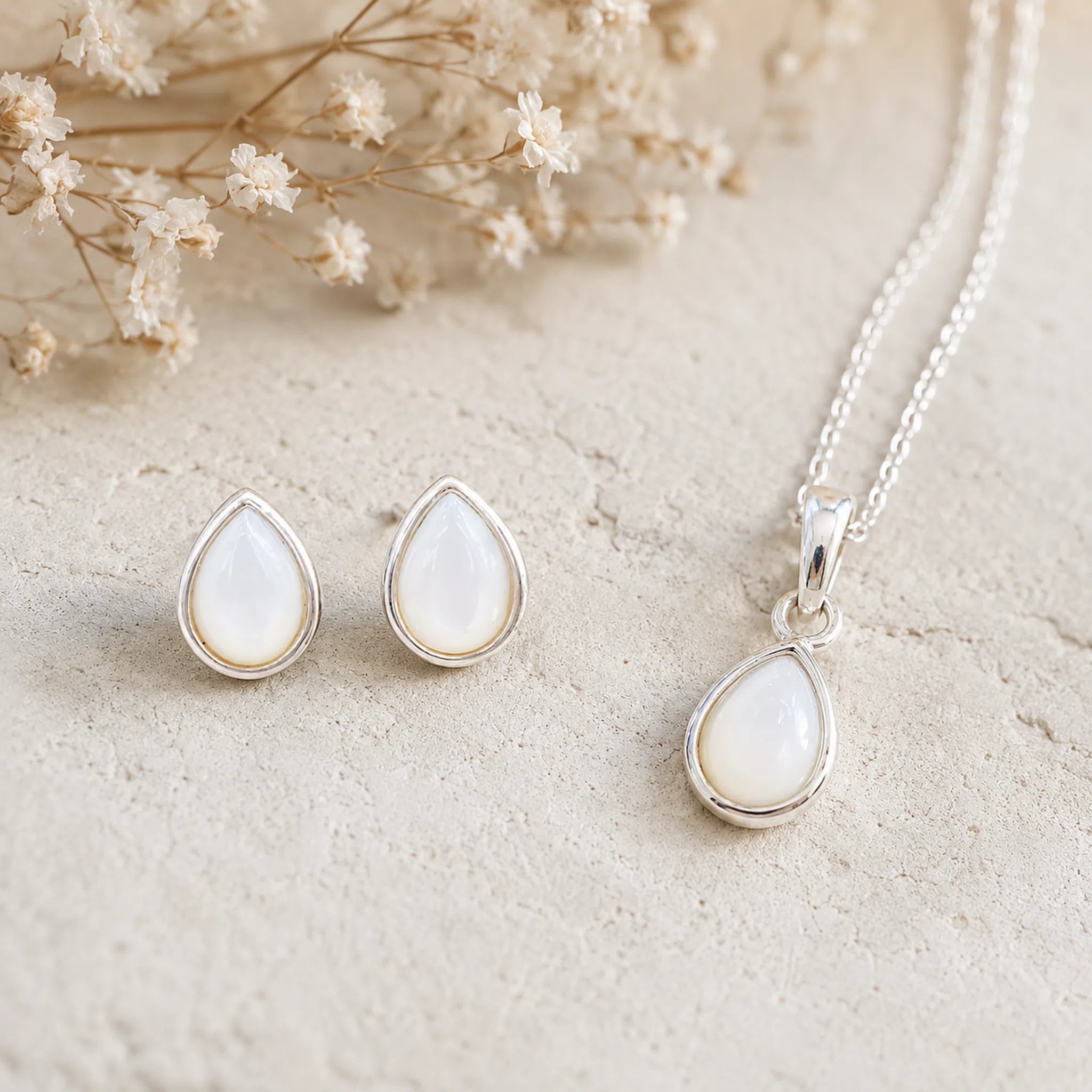 Sterling Silver June Birthstone Jewellery set featuring a teardrop pendant pearl necklace and matching stud pearl earrings with natural milky white teardrop pearls, arranged on a textured beige surface with dried flowers.