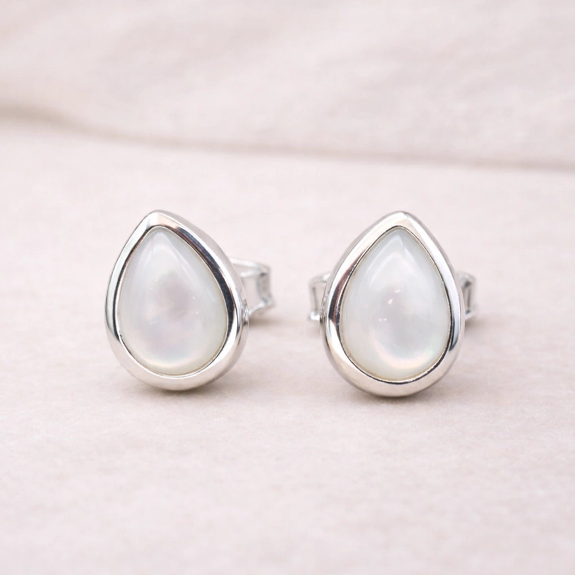 Teardrop-shaped sterling silver stud earrings with a light-colored natural pearl gemstone on a soft pink background