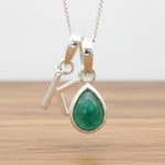 Silver Initial K necklace with a teardrop-shaped Emerald gemstone pendant hanging above a rustic wooden surface