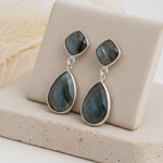 Teardrop-shaped labradorite earrings on a beige stand with a soft white background