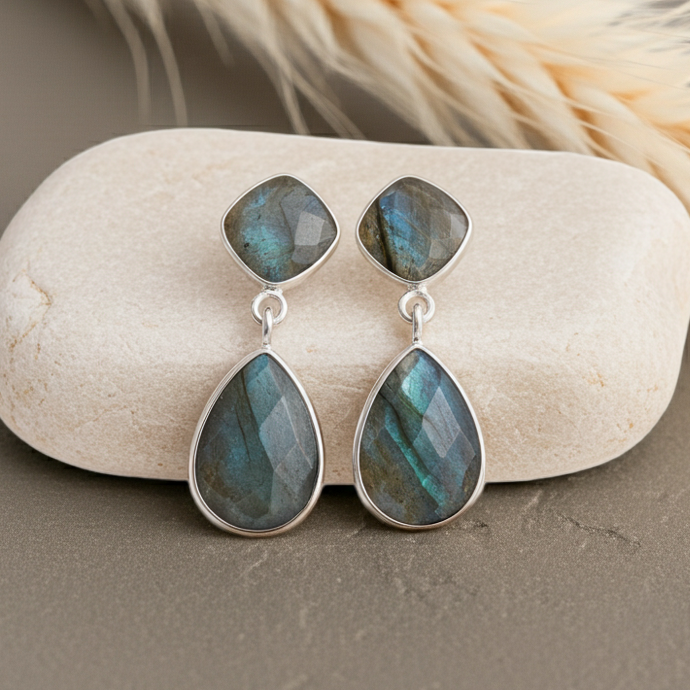 Teardrop-shaped earrings with labradorite stones on a stone surface with dried grass in the background.