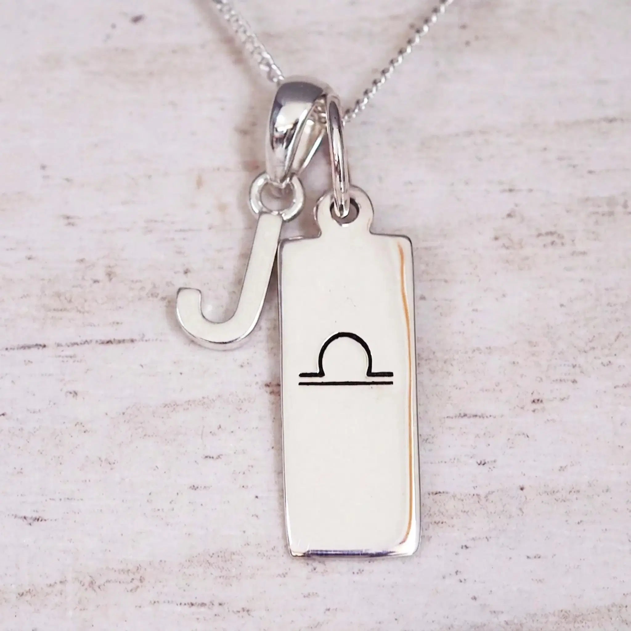 Silver Initial and Libra Zodiac Necklace with a letter J pendant and engraved Libra bar, flat lay on light wood