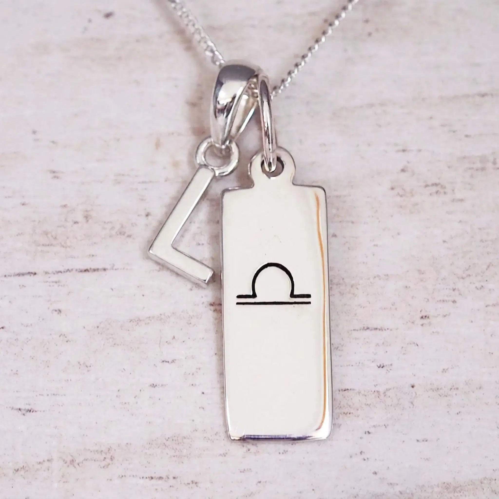 Silver Initial and Libra Zodiac Necklace with a letter L pendant and engraved Libra bar, flat lay on light wood