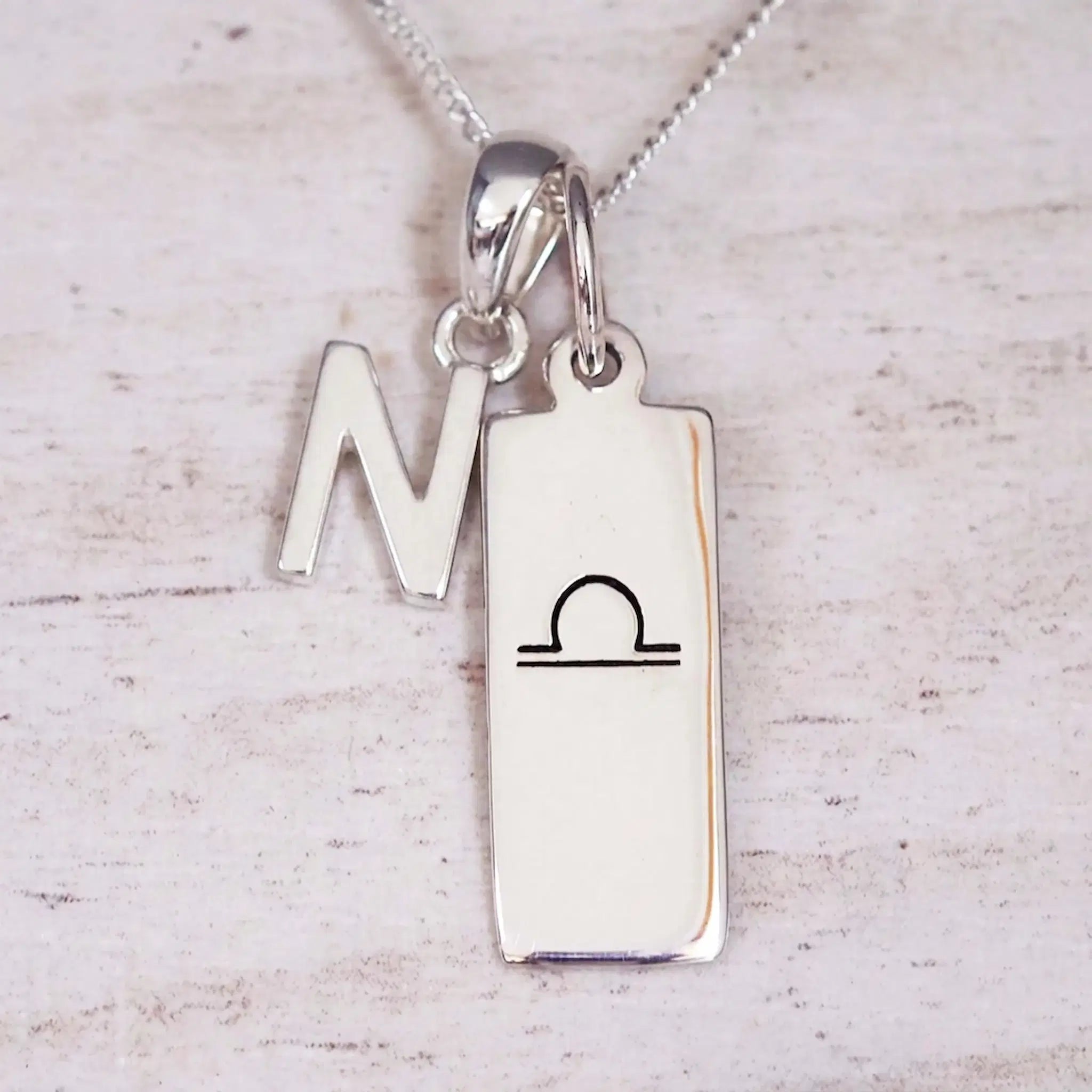 Silver Initial and Libra Zodiac Necklace with a letter N pendant and engraved Libra bar, flat lay on light wood