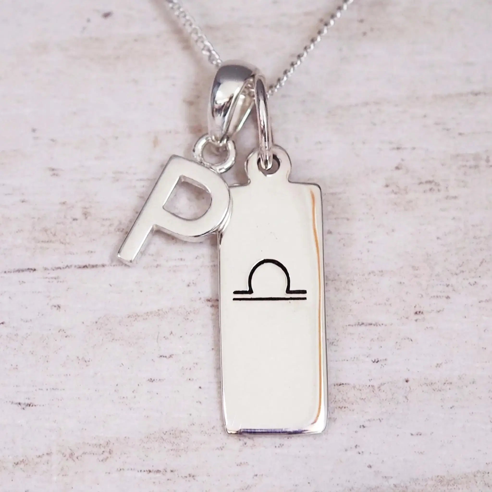 Silver Initial and Libra Zodiac Necklace with a letter P pendant and engraved Libra bar, flat lay on light wood