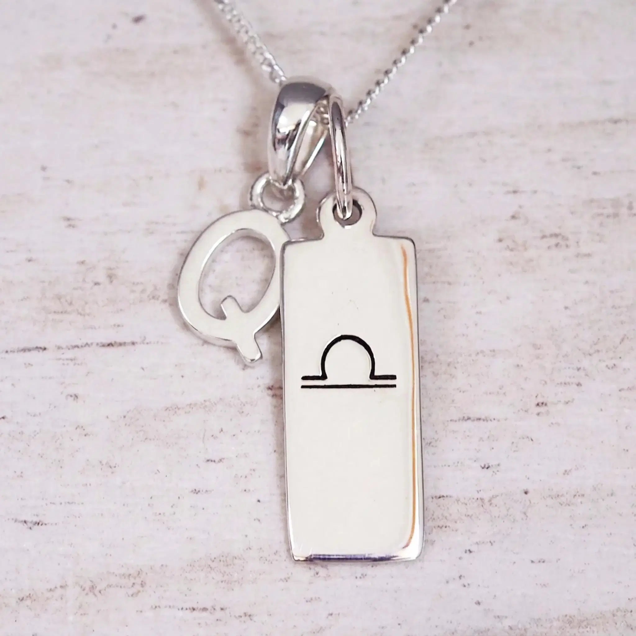 Silver Initial and Libra Zodiac Necklace with a letter Q pendant and engraved Libra bar, flat lay on light wood