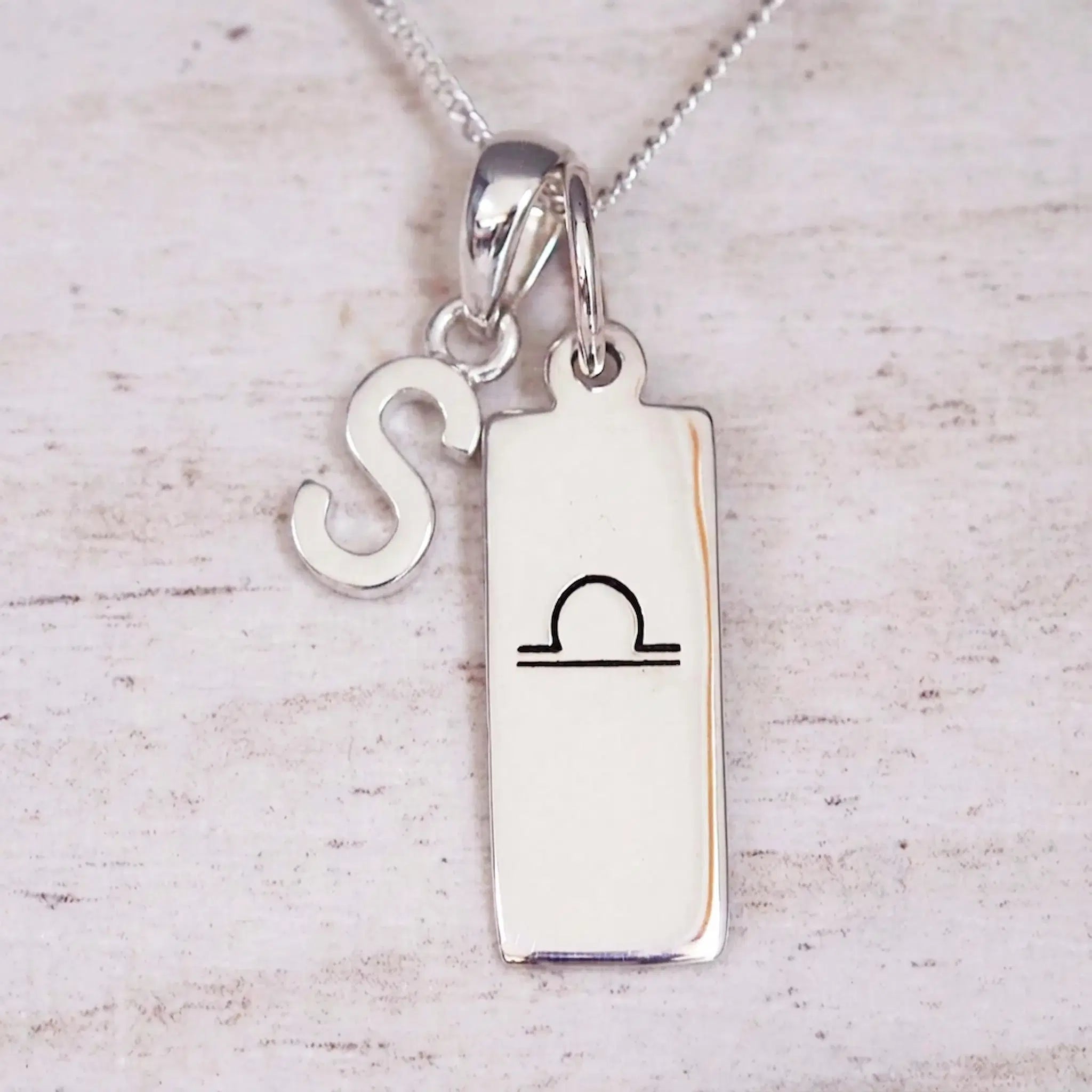 Silver Initial and Libra Zodiac Necklace with a letter S pendant and engraved Libra bar, flat lay on light wood