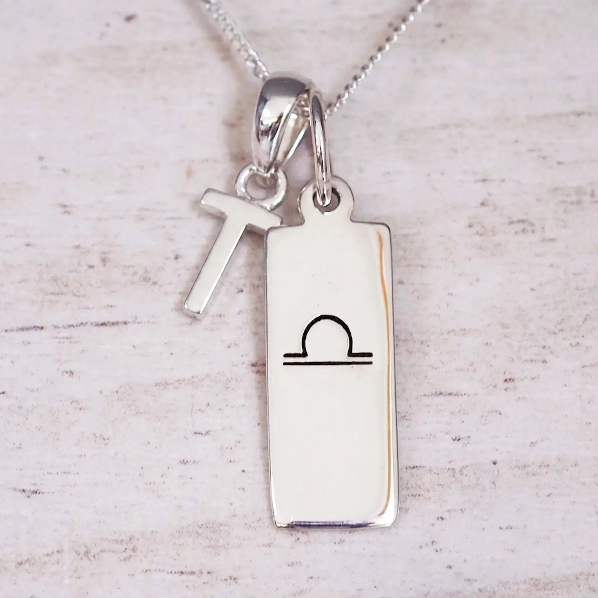 Silver Initial and Libra Zodiac Necklace with a letter T pendant and engraved Libra bar, flat lay on light wood
