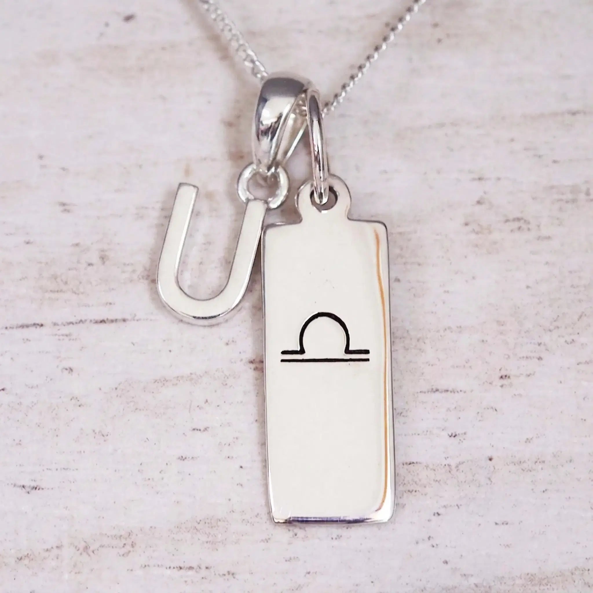 Silver Initial and Libra Zodiac Necklace with a letter U pendant and engraved Libra bar, flat lay on light wood
