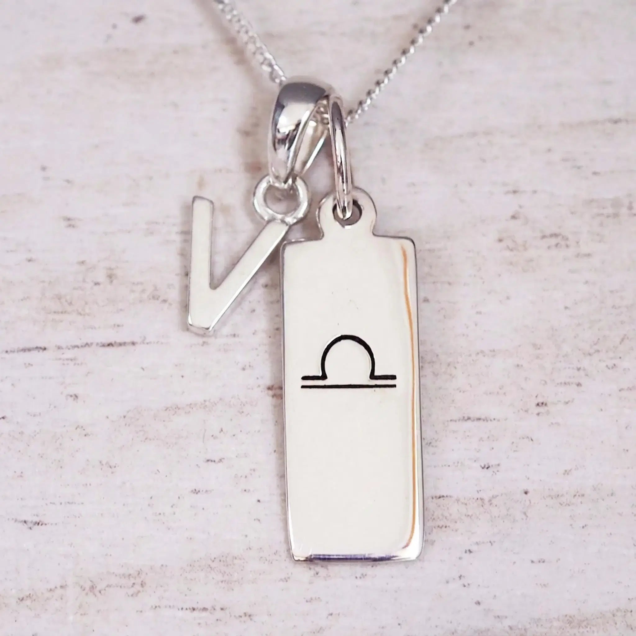 Silver Initial and Libra Zodiac Necklace with a letter V pendant and engraved Libra bar, flat lay on light wood