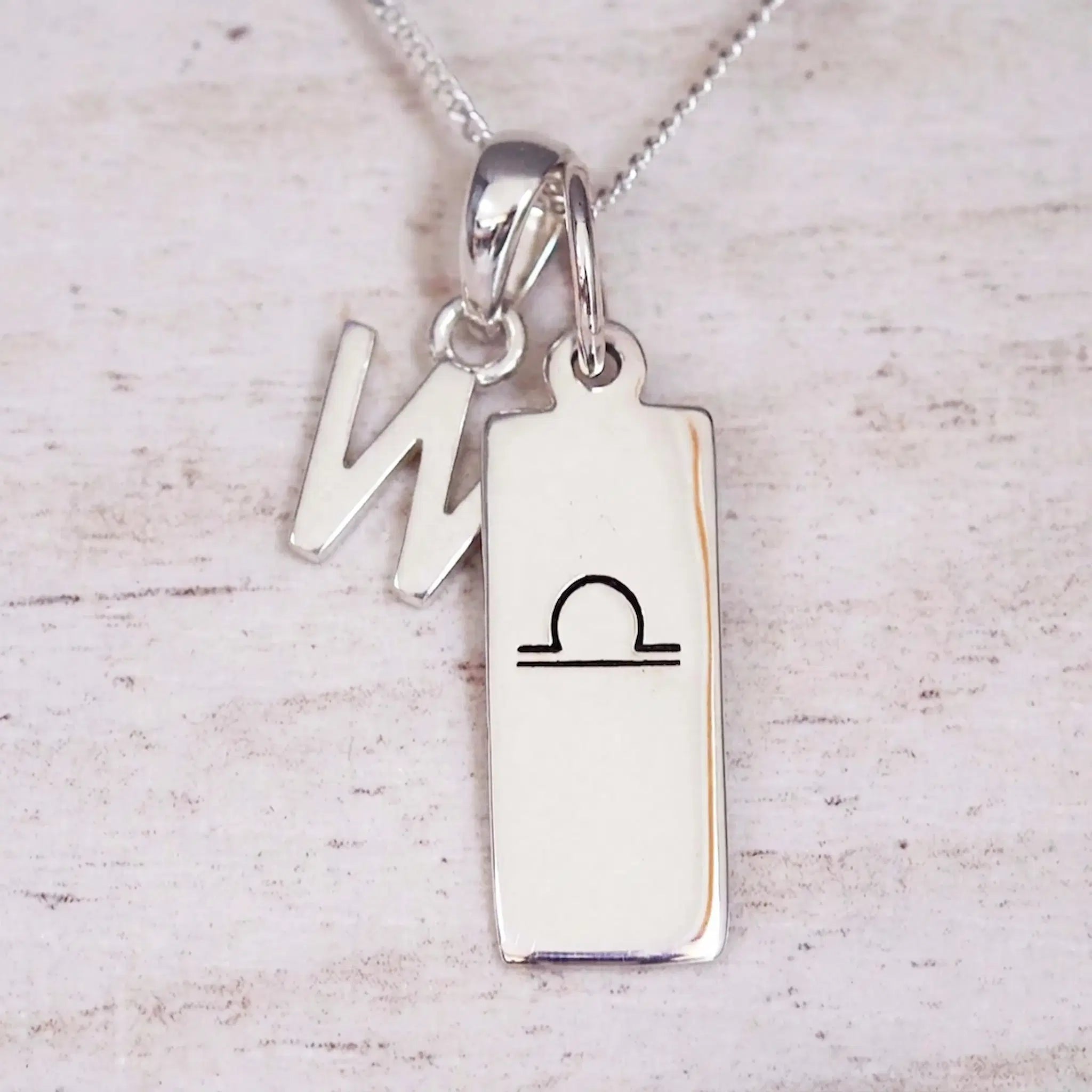 Silver Initial and Libra Zodiac Necklace with a letter W pendant and engraved Libra bar, flat lay on light wood