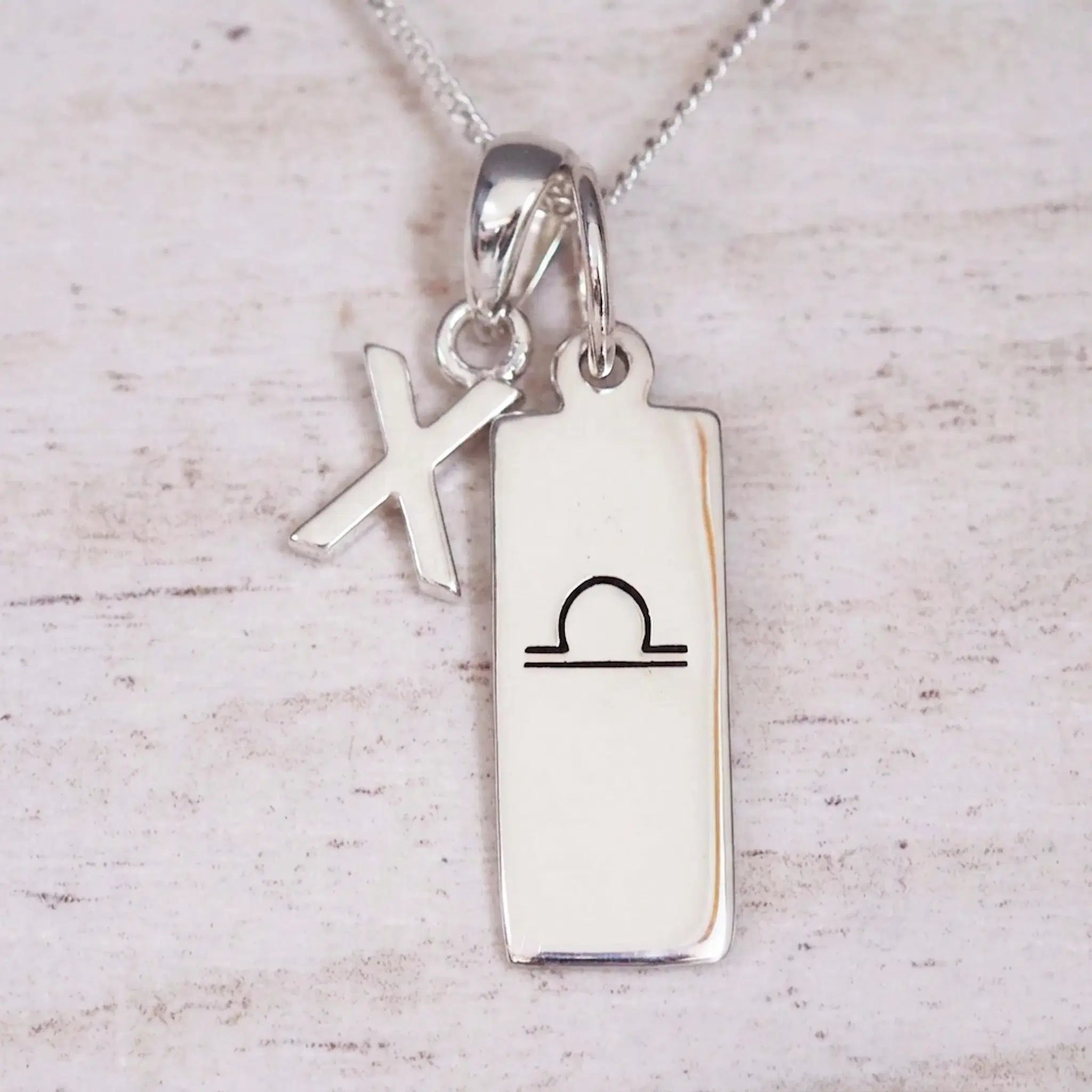 Silver Initial and Libra Zodiac Necklace with a letter X pendant and engraved Libra bar, flat lay on light wood