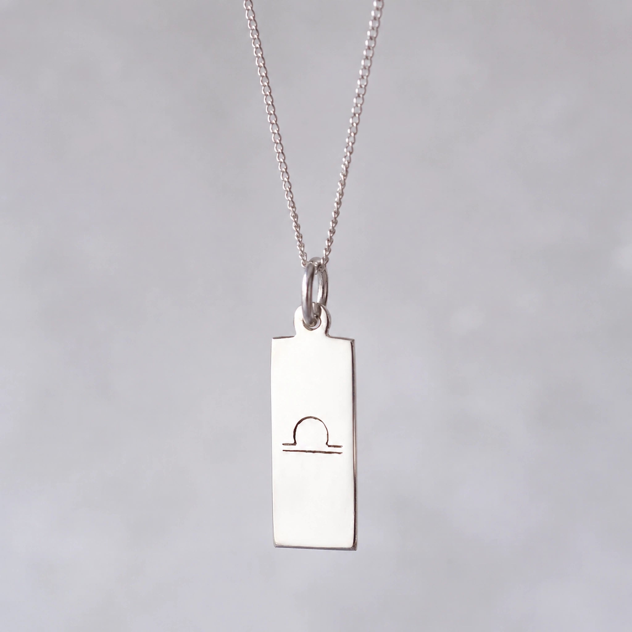 Silver necklace with a rectangular libra zodiac glyph pendant on a grey background