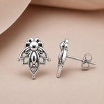 A pair of silver stud earrings with an intricate floral or leaf-like cutout design, shown on a soft beige background. One earring is facing forward to show the front design, and the other is turned sideways to show the post and butterfly back.