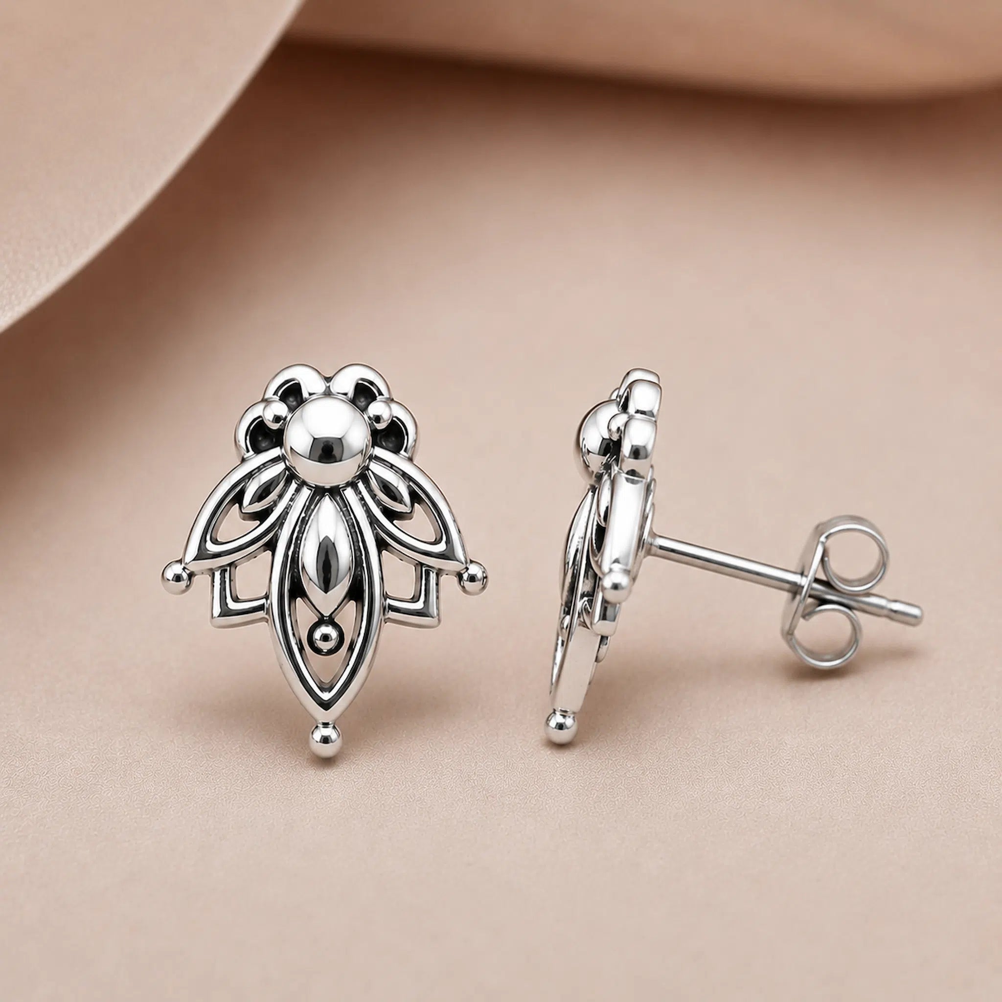 A pair of silver stud earrings with an intricate floral or leaf-like cutout design, shown on a soft beige background. One earring is facing forward to show the front design, and the other is turned sideways to show the post and butterfly back.