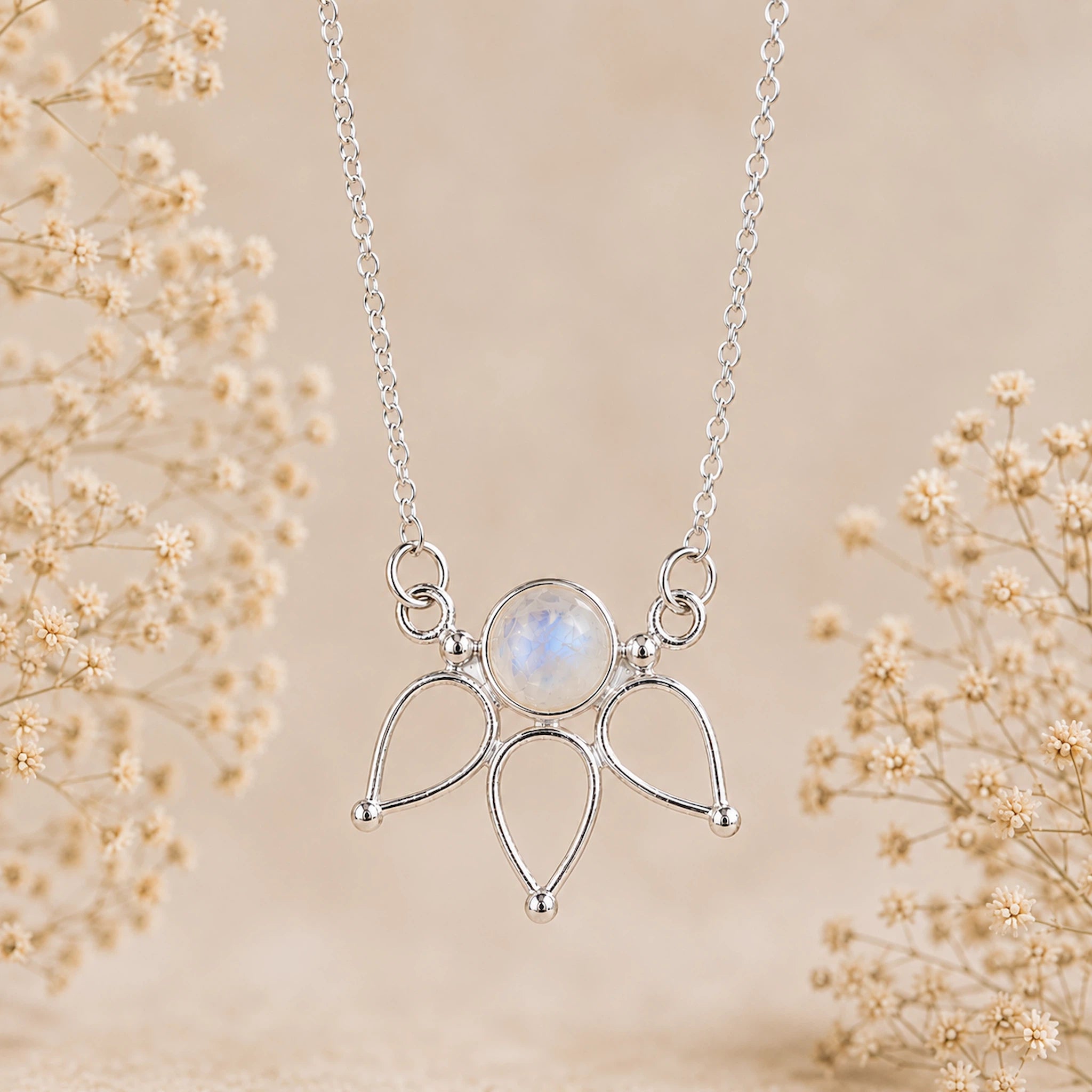 A silver pendant moonstone necklace featuring a central round iridescent moonstone surrounded by three teardrop-shaped metal settings, suspended from a delicate chain. The necklace is displayed against a soft beige background decorated with dried baby's breath flowers.