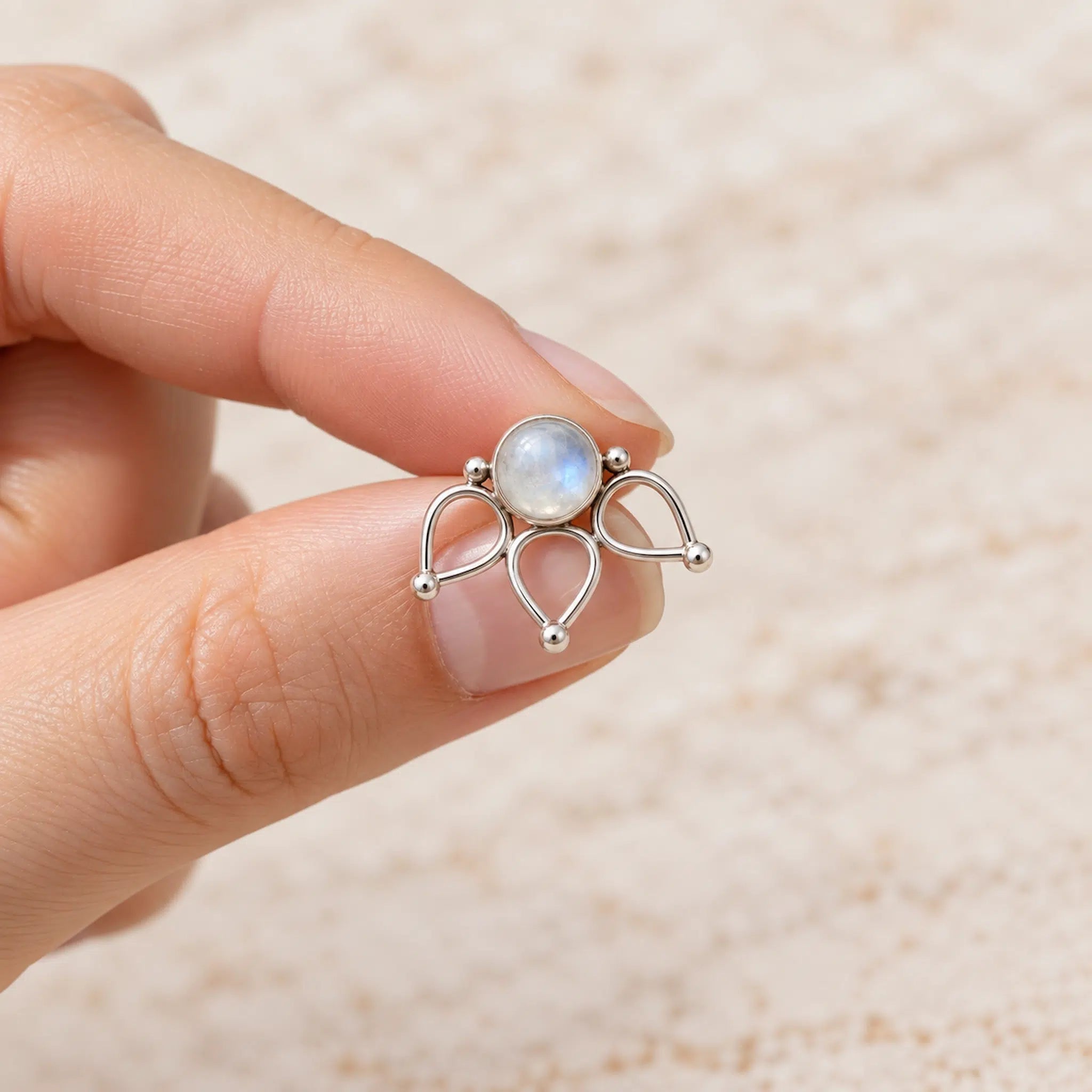 A silver lotus flower stud earring with a round iridescent moonstone center, held between a person's fingers against a soft beige textured background.
