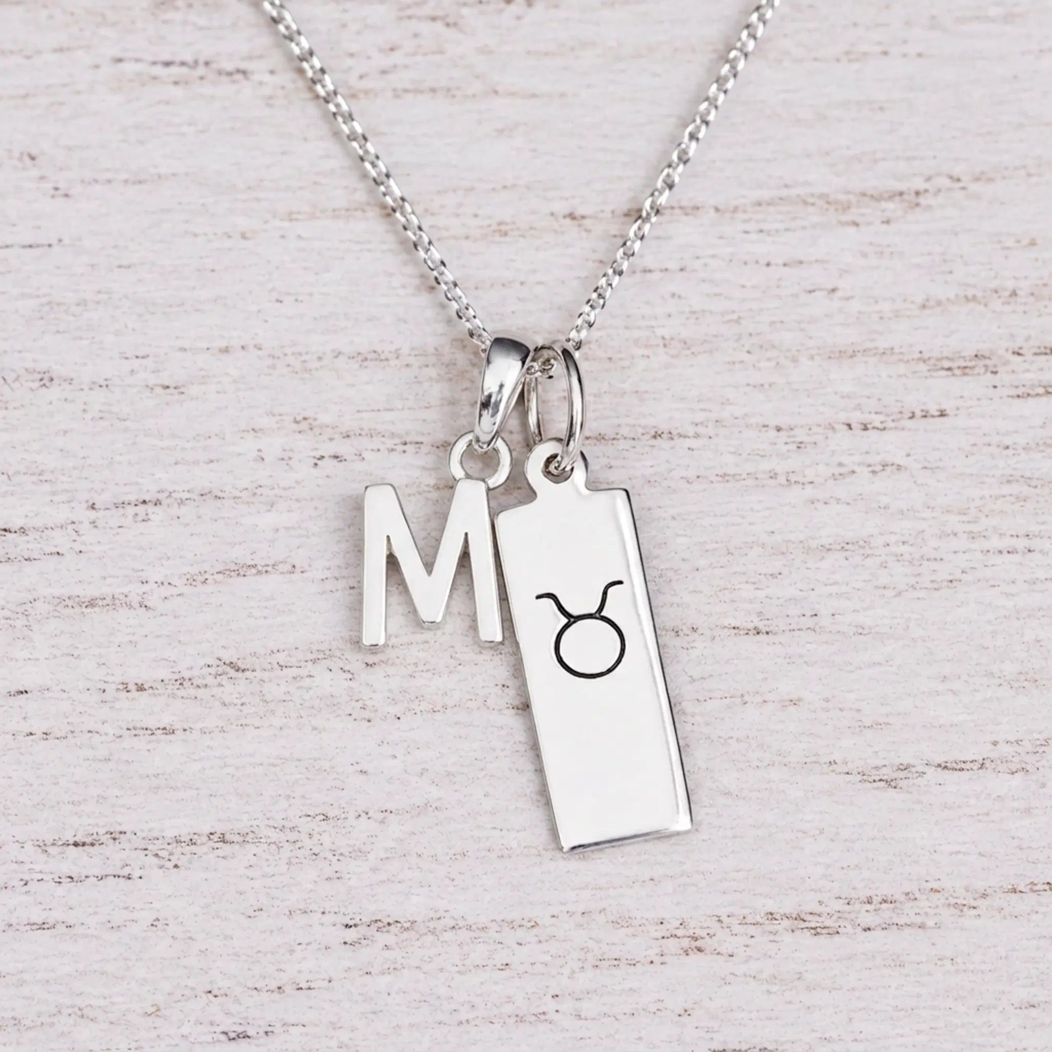 Sterling silver M initial Pendant and Taurus Necklace on a fine silver chain on a light wooden background
