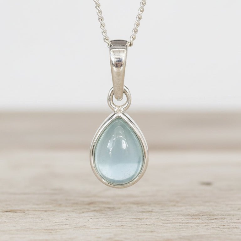 Teardrop-shaped pendant with light blue aquamarine gemstone and silver frame on a neutral background