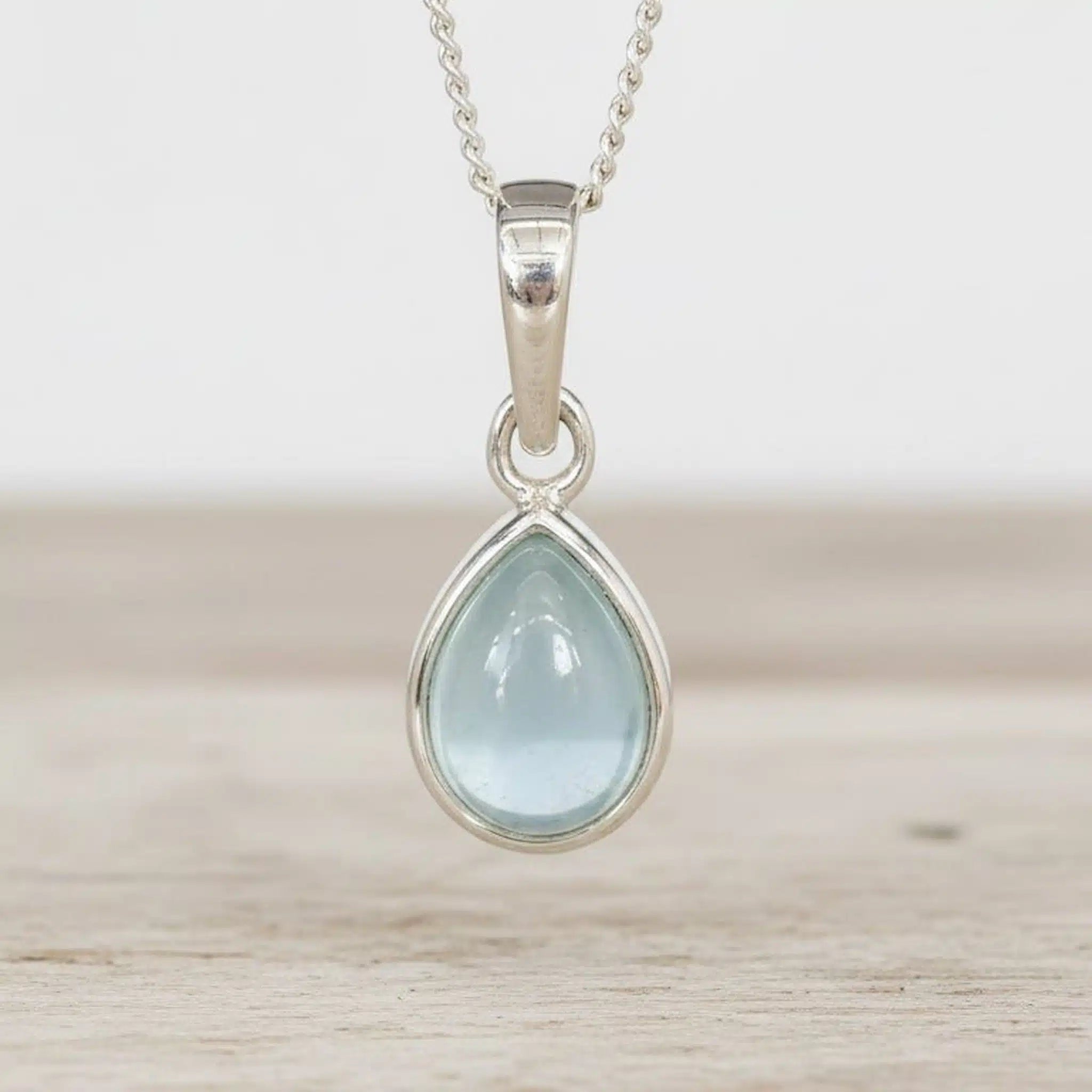 march birthstone necklace Teardrop-shaped natural aquamarine gemstone pendant on a sterling silver chain against a wooden background