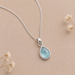 A silver march birthstone necklace with teardrop pendant necklace with a light blue natural aquamarine gemstone, displayed on a beige background with small dried flowers.