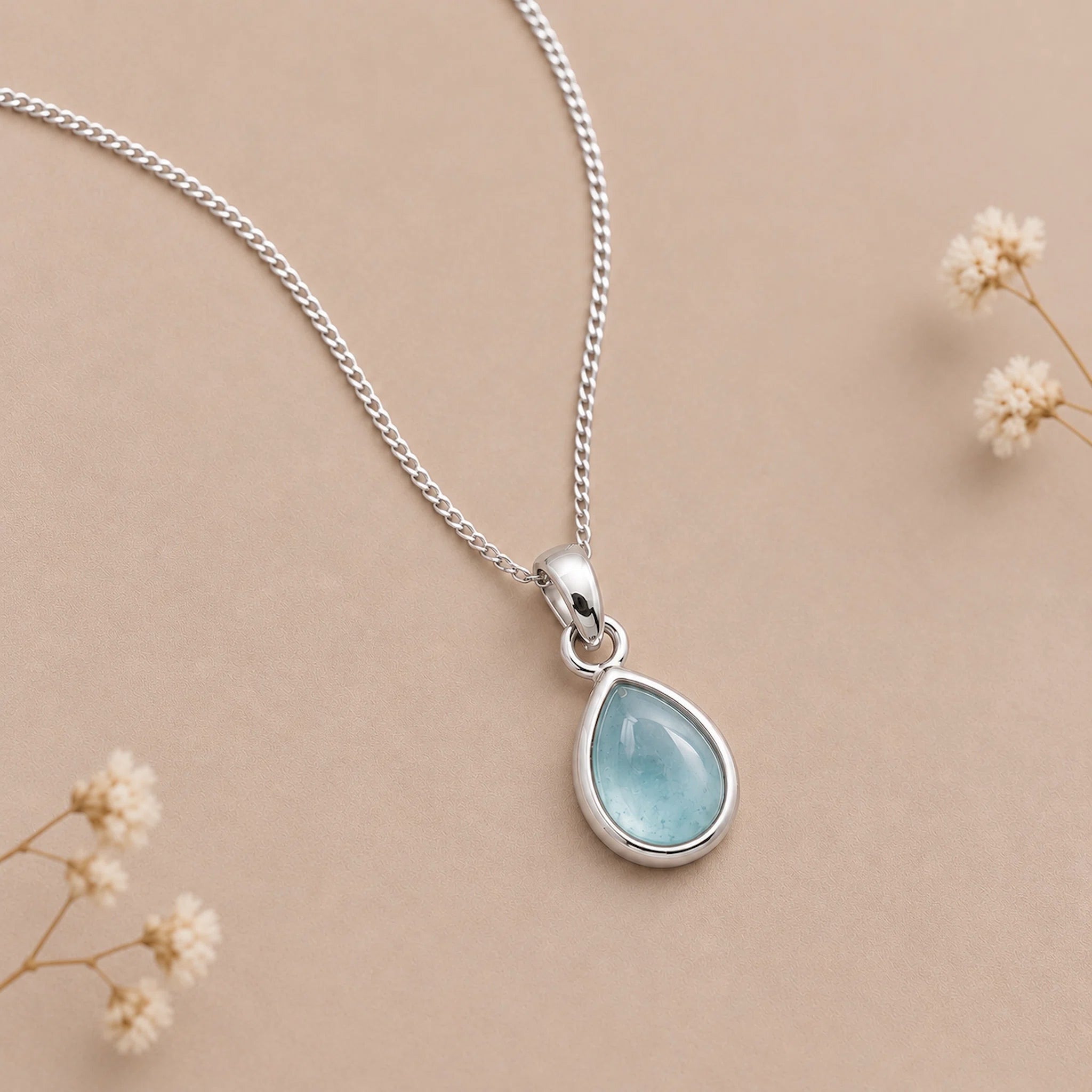 A silver march birthstone necklace with teardrop pendant necklace with a light blue natural aquamarine gemstone, displayed on a beige background with small dried flowers.