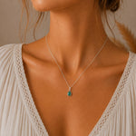A close-up of a woman wearing a delicate silver chain may birthstone necklace with a small teardrop-shaped natural emerald green pendant, styled with a cream-colored lace top.