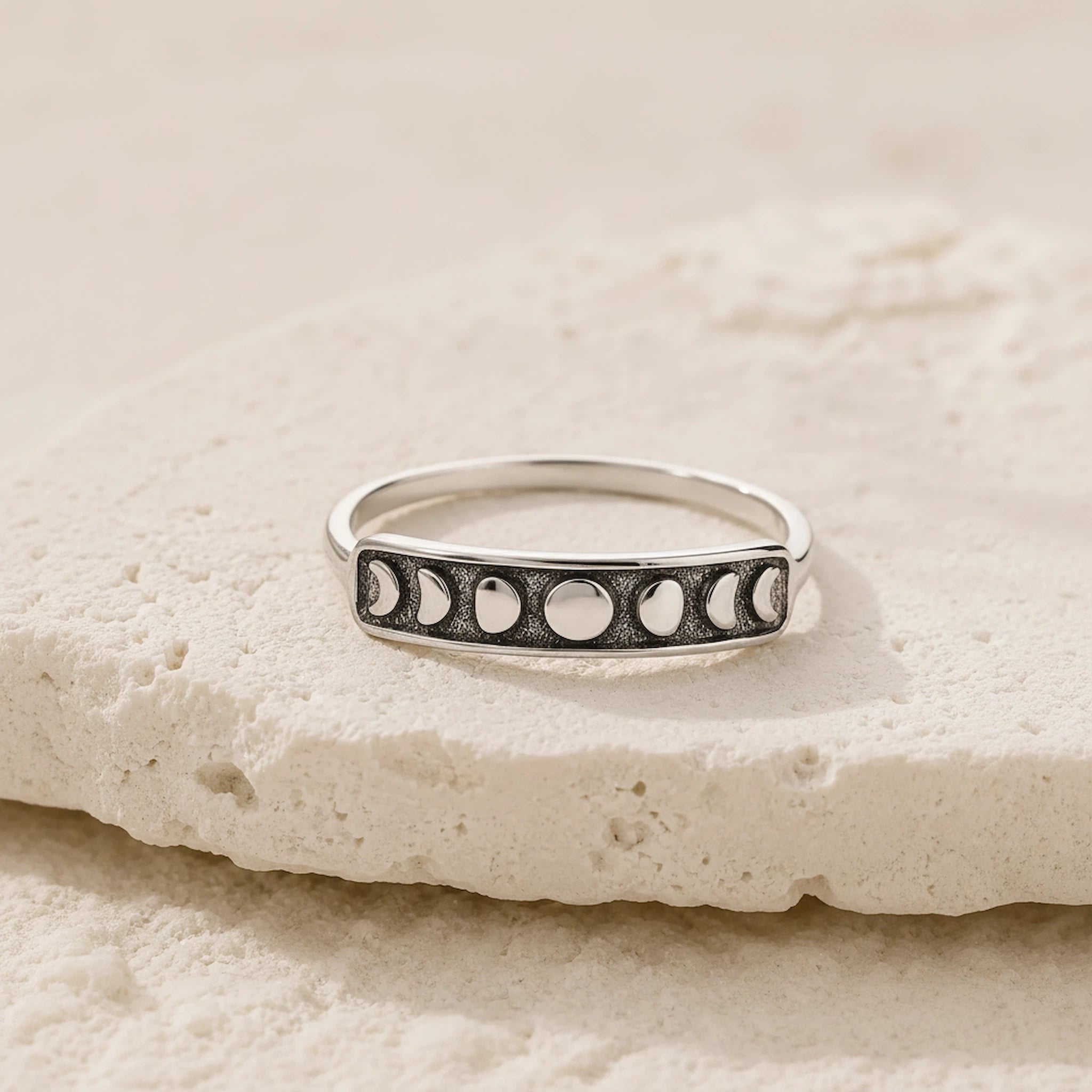 A silver band ring featuring a row of moon phase motifs, resting on a textured beige stone slab.