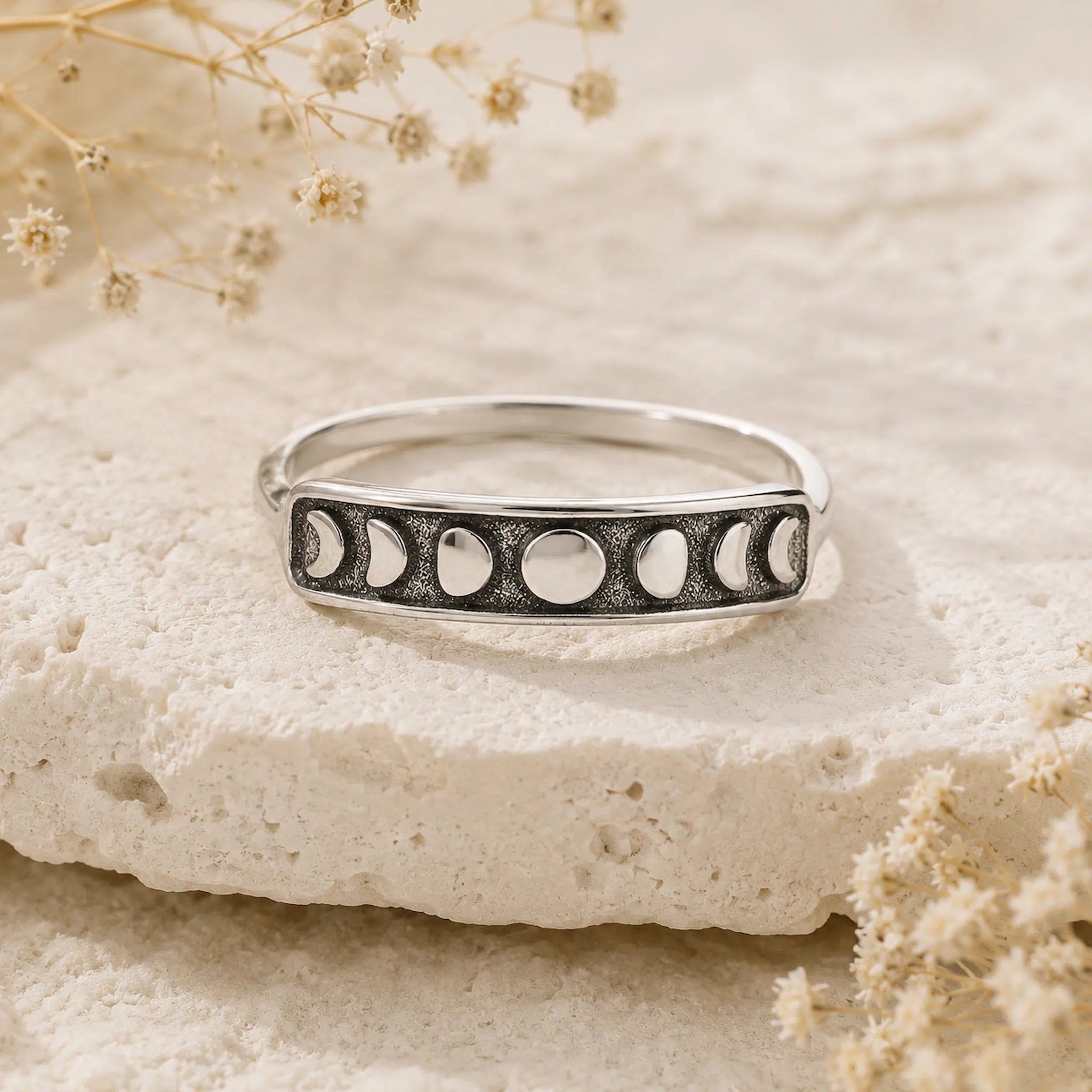 A silver ring featuring a moon phase design, resting on a textured beige stone slab with dried flowers in the background.