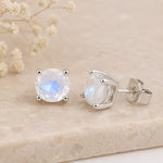 A pair of oval-cut moonstone stud earrings in silver prong settings, displayed on a light beige stone surface with dried baby's breath flowers in the background.