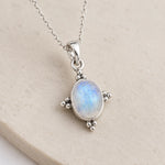 A silver pendant necklace featuring an oval iridescent moonstone with blue and white flashes, set in a four-prong silver bezel on a matching chain, displayed on a textured beige stone surface.
