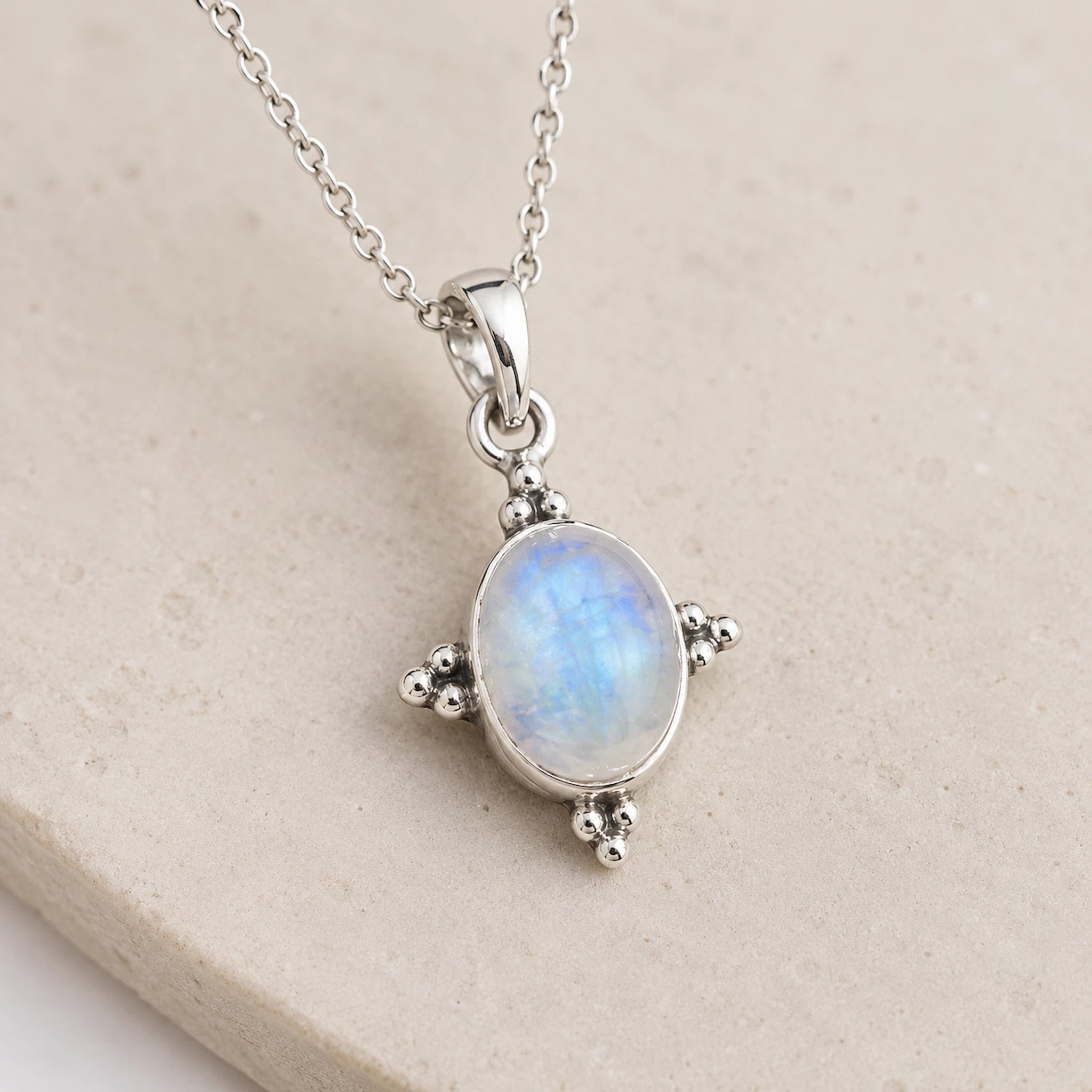 A silver pendant necklace featuring an oval iridescent moonstone with blue and white flashes, set in a four-prong silver bezel on a matching chain, displayed on a textured beige stone surface.
