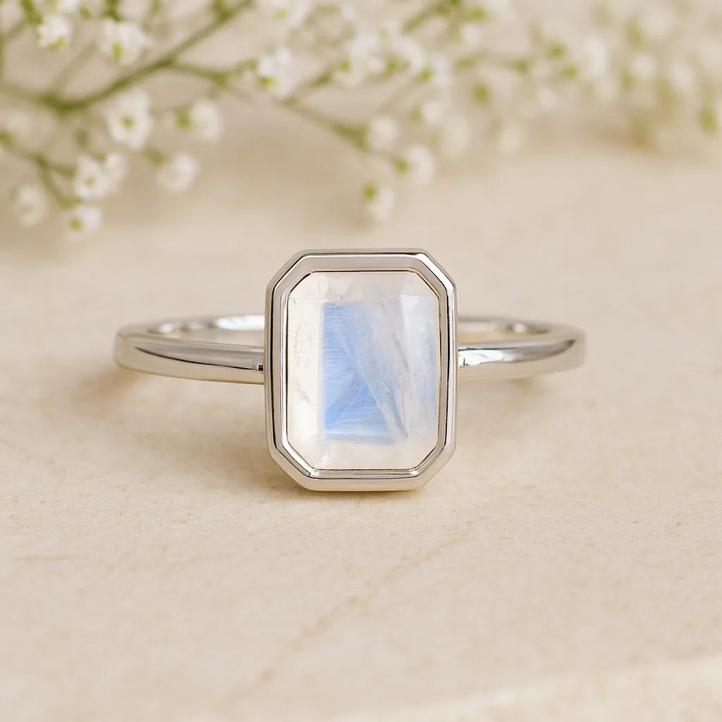 Silver ring with a rectangular moonstone stone on a beige surface with blurred flowers in the background