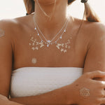 A woman outdoors wearing a silver necklace with natural moonstone, delicate silver floral temporary tattoos on her chest, shoulders, and wrist, with a white strapless top and blurred hills in the background.