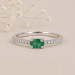 A silver emerald ring featuring an oval-cut natural green emerald gemstone in a four-prong setting, with a pavé band of small clear stones, photographed on a warm beige background with delicate dried flowers.
