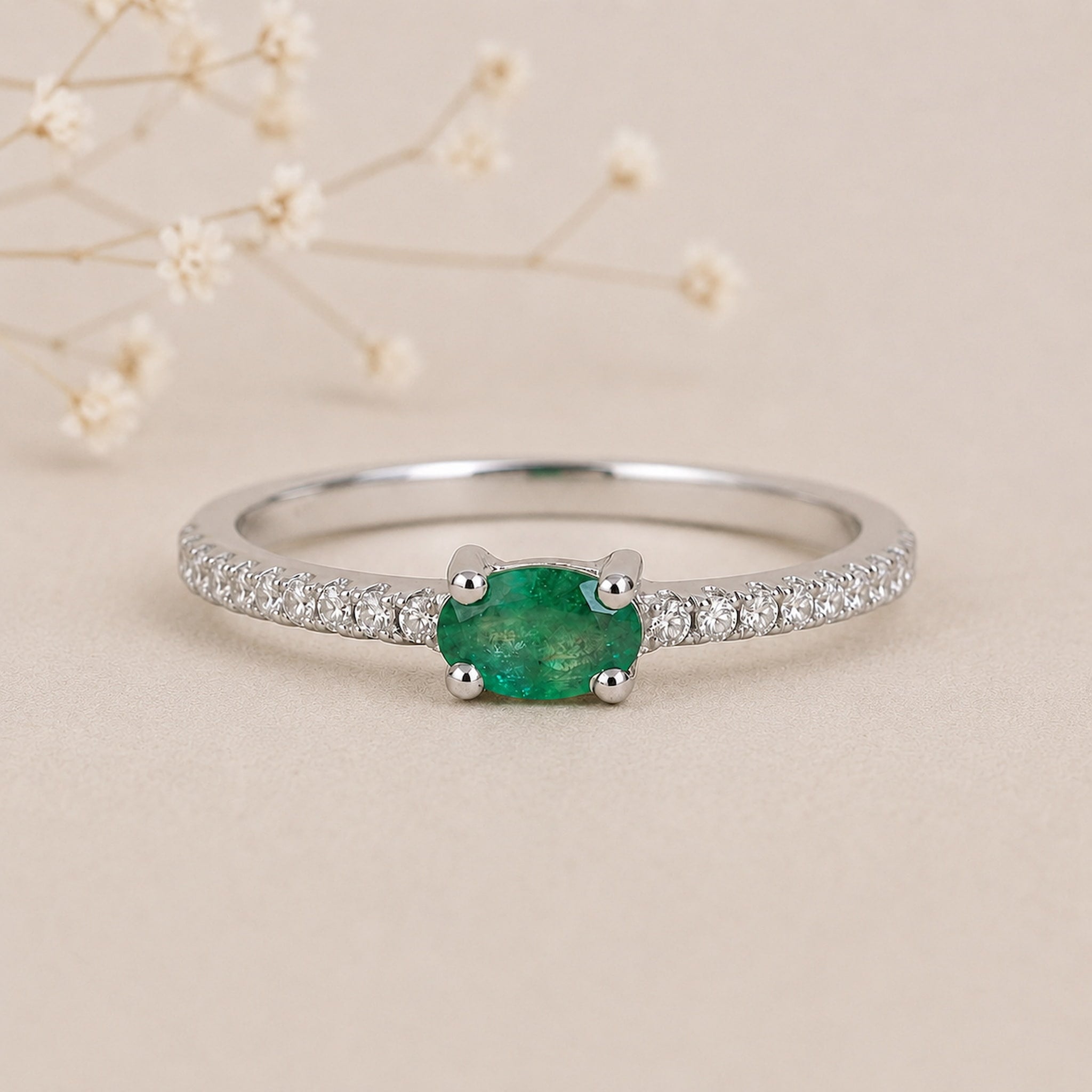 A silver emerald ring featuring an oval-cut natural green emerald gemstone in a four-prong setting, with a pavé band of small clear stones, photographed on a warm beige background with delicate dried flowers.