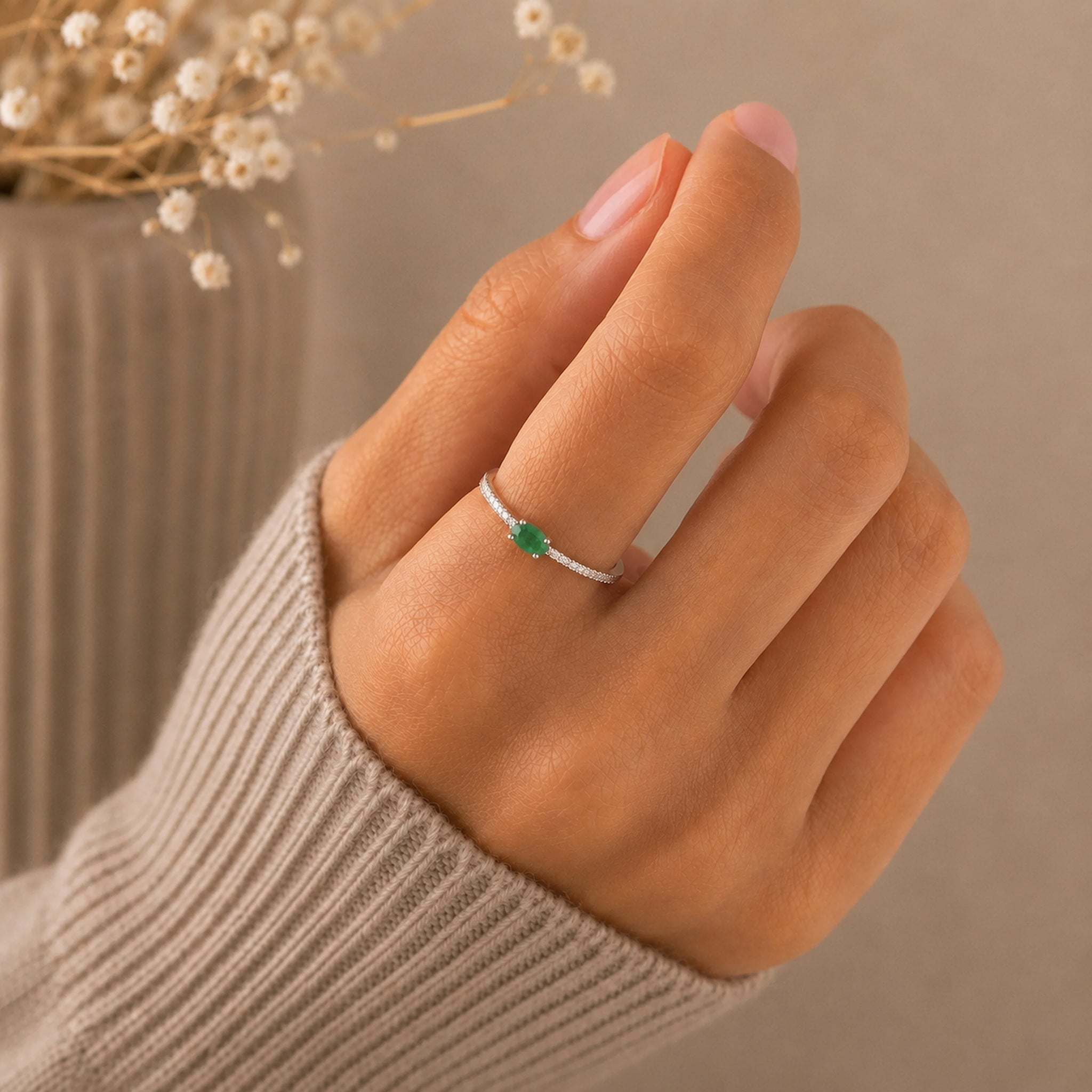A close-up of a hand wearing a delicate silver ring with a small faceted natural emerald green gemstone, styled with a beige ribbed sweater sleeve against a soft neutral background.